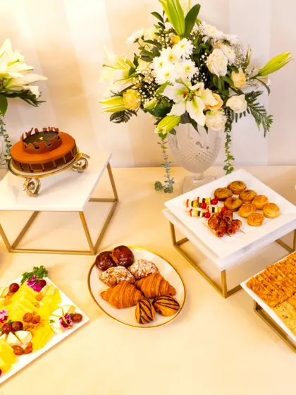 A bright and cheerful high-tea setup featuring a beautiful cake, fresh croissants, and a colorful cheese platter. The display is accented with stunning white lily and daisy floral arrangements, perfect for a celebratory brunch or afternoon event.