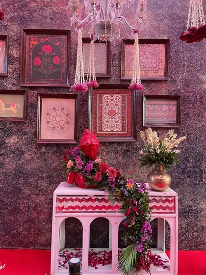 A detail shot from the red and gold Shreenathji wedding, showing a beautifully decorated corner. A console with traditional arches is adorned with a floral runner and framed artwork, showcasing how every element is curated to fit the grand theme.