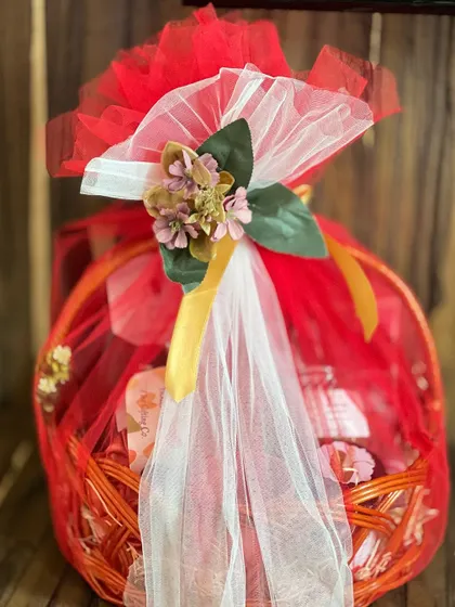A vibrant corporate farewell gift hamper. The bright red basket is wrapped in red and white net with a floral topper, creating a cheerful and memorable goodbye gift.