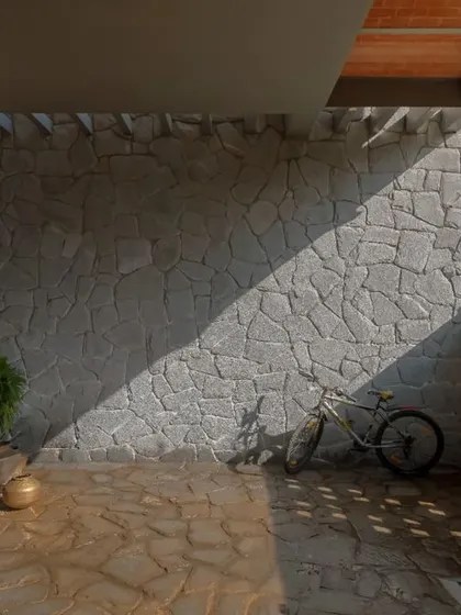 The entrance yard features a Sira stone wall and floor laid in a random rubble pattern. The diagonal shadow cutting across the textured surfaces sets the tone for the earthy, tactile experience of the home.