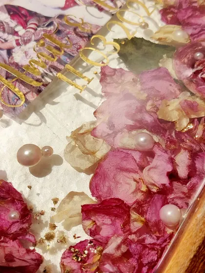 A close-up on the details of a teakwood frame preservation. The delicate pearls and gold text stand out against the soft pink petals, creating a look of pure elegance.