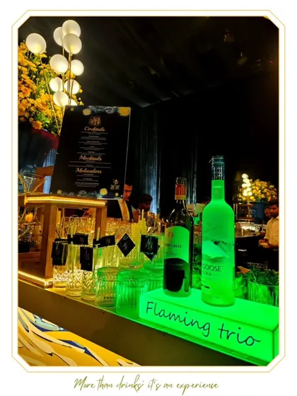 A clean and modern bar setup with green LED under-lighting and premium spirits on display. The prominent "Flaming Trio" branding adds a professional touch.