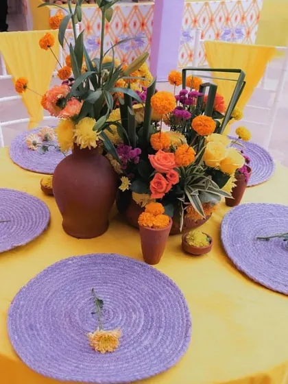 A close-up of a table setting for a Haldi lunch. The decor features a yellow tablecloth with purple placemats and a centerpiece of marigolds and roses.