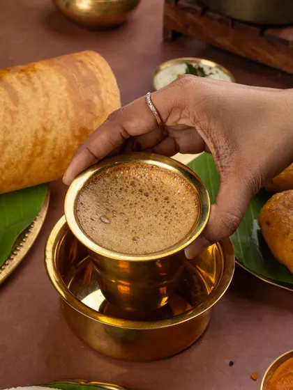 A hand holding a traditional brass cup of filter coffee, completing the authentic South Indian breakfast experience.