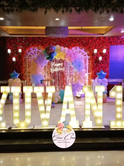 A grand stage setup for a first birthday with large marquee letters spelling 'YUVAN 1' and a pastel balloon arch.