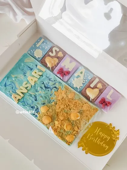 A custom beach-themed chocolate bar and smaller initial chocolates, made for a special birthday. The wave effect and biscuit "sand" create a fun, edible scene.
