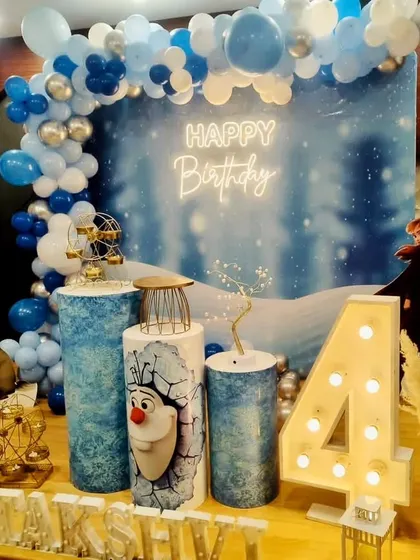 This Frozen-themed 4th birthday party includes a snowy forest backdrop, an Olaf-themed pedestal, and a light-up marquee number.