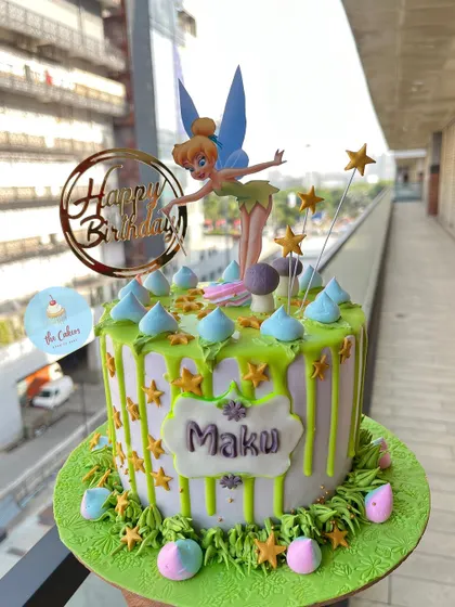 A magical Tinker Bell cake with a green drip design. The cake is adorned with a Tinker Bell figure, golden stars, and buttercream grass, creating a whimsical scene from Pixie Hollow.