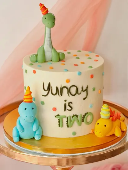 A colourful dinosaur party cake for a second birthday. This design features three friendly fondant dinosaurs wearing party hats on a polka-dotted buttercream base.