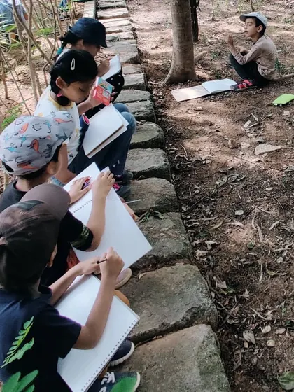 Children sit along a stone path, deeply engaged in sketching and writing in their nature journals during a park visit.