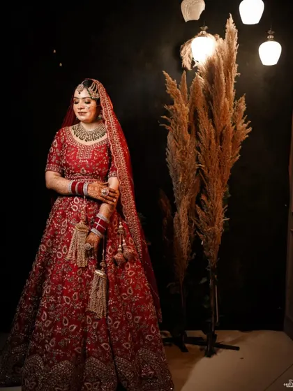 A classic bridal portrait. The bride stands elegantly, her red lehenga and intricate jewelry shining against the dark, rustic backdrop.
