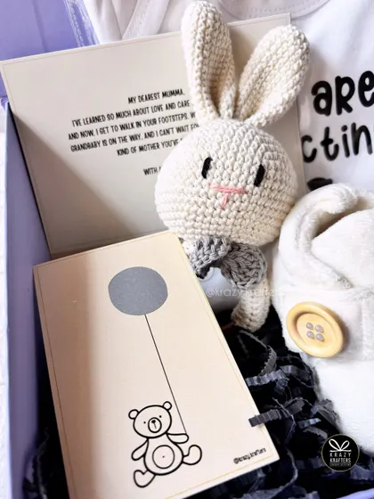 A complete view of our scratch-card announcement hamper, including a personalized message card to the new grandma, a onesie, and a cute bunny rattle.