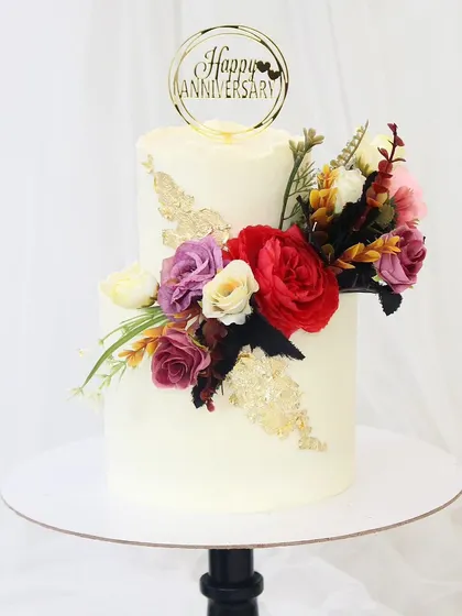 A classic and beautiful anniversary cake. This two-tiered design features clean white buttercream, gold leaf accents, and a stunning arrangement of fresh, colorful flowers to celebrate lasting love.