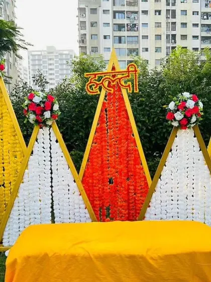 A modern geometric Haldi backdrop using triangular frames decorated with white and orange flower strings. This is a contemporary take on traditional floral decorations.