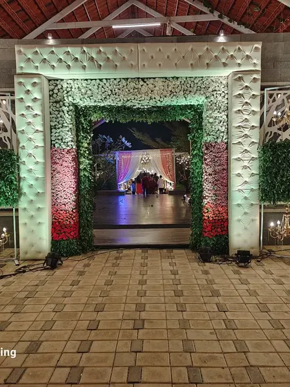 A grand main entrance designed for a wedding reception. The archway is covered in a mix of white, pink, and red flowers, framed by lush greenery and elegant white cushioned panels.