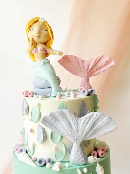 A two-tiered mermaid cake with a hand-sculpted mermaid figure. The design features pastel colours and detailed fondant mermaid tails.
