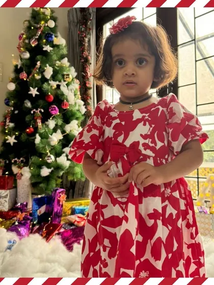 A toddler stands by the Christmas tree, dressed beautifully for the celebration.