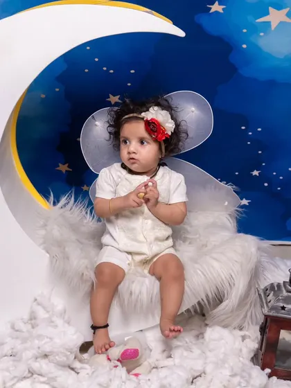 A dreamy 'on the moon' setup with a starry night backdrop and fluffy clouds. The baby is dressed as a little angel, sitting on a crescent moon prop.