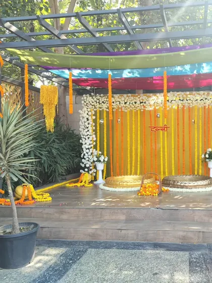 An outdoor Haldi setup on a wooden deck. The backdrop of marigold strings is framed by white flowers, and the two urlis are ready for the ceremony, creating a beautiful contrast with the natural surroundings.