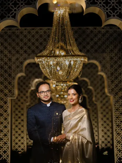 A classic and elegant reception portrait. The couple stands before a beautifully lit backdrop featuring intricate latticework and a stunning crystal chandelier.