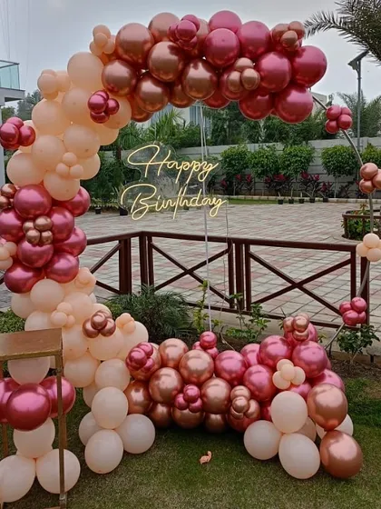 An outdoor birthday decoration with a balloon arch in peach and metallic pink, complete with a neon sign.