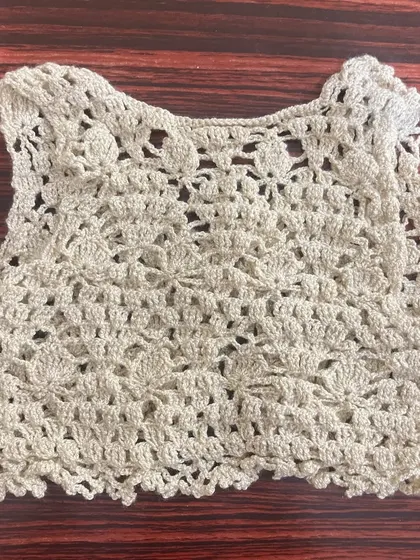 A delicate, lacy crochet crop top or vest in a neutral beige color, perfect for layering over a baby's outfit in milder weather.