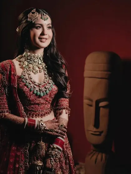 A stunning portrait of the bride in her red lehenga, her regal look enhanced by layered polki and emerald necklaces, embodying the grace of a modern maharani.