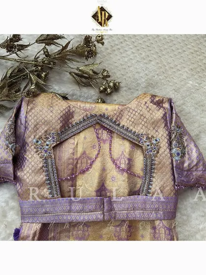 The back of a lavender brocade blouse, featuring a unique cutout design framed with intricate band embroidery. The saree's own zari is used to create a matching belt.