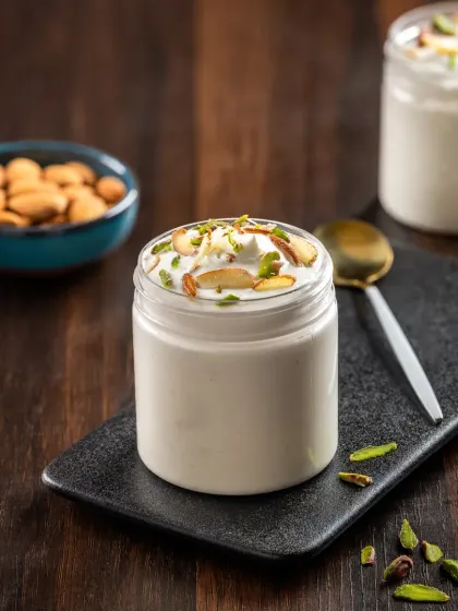 A creamy white jar dessert topped with chopped pistachios and almonds, styled simply to emphasize its texture and ingredients.