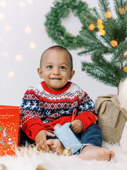 A baby boy in a festive sweater, smiling brightly next to the Christmas tree. His joy is infectious.
