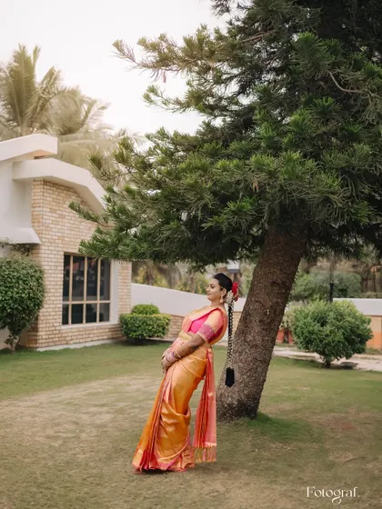 A serene portrait of a South Indian bride in a beautiful orange saree, leaning against a tree in a peaceful garden setting.