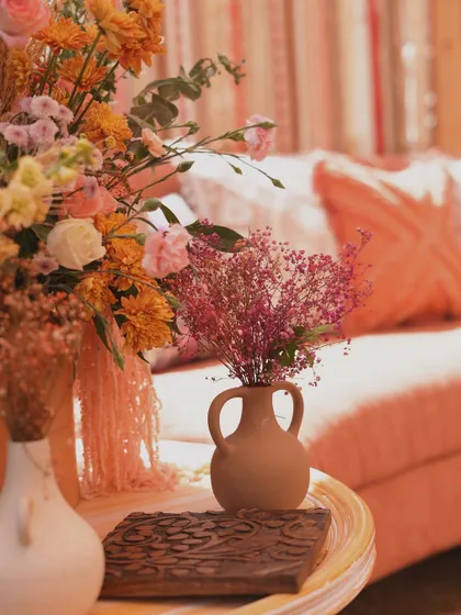 A detail shot of a rustic terracotta vase holding delicate pink flowers, placed on a table with a carved wooden block.
