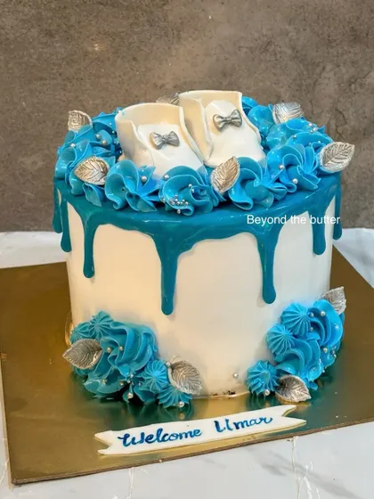 It's a boy! This welcome cake for baby Umar features a beautiful blue drip, blue buttercream rosettes, and tiny, handcrafted white fondant baby shoes.