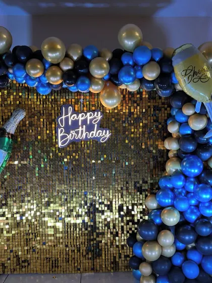 A vibrant birthday party backdrop with a gold shimmer wall. The decor includes a blue, black, and gold balloon arch and champagne-themed foil balloons.