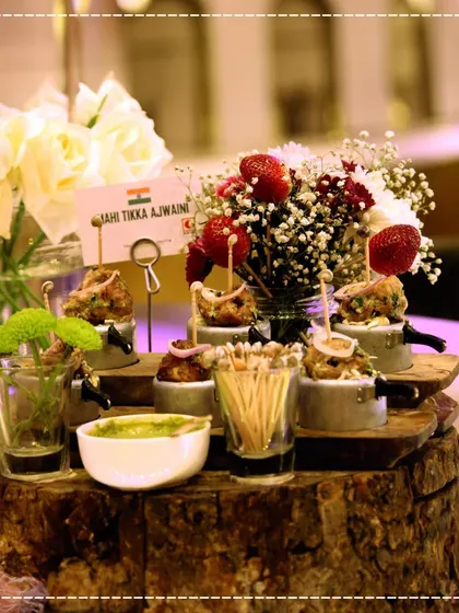 An appetizer station featuring Mahi Tikka Ajwaini, served on miniature pressure cookers set on a rustic wooden log, a creative and memorable presentation.