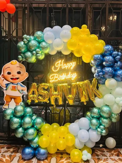A Cocomelon themed birthday photo booth with a circular balloon frame. The setup includes a neon 'Happy Birthday' sign and a cutout of a Cocomelon character, perfect for pictures.