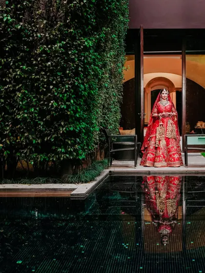 A stunning portrait of the bride by the pool, her reflection captured in the dark water. The contrast between her red lehenga and the green foliage is striking.