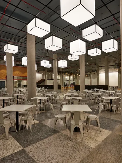 An industrial-style cafeteria with a checkerboard terrazzo floor, concrete pillars, and large, suspended box lights. The design is raw, spacious, and highly durable.