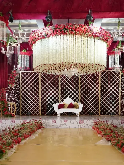 A magnificent reception stage featuring a massive cylindrical chandelier of white floral strings, set against a backdrop of red velvet and golden lattice. This design is the epitome of opulence and grandeur.