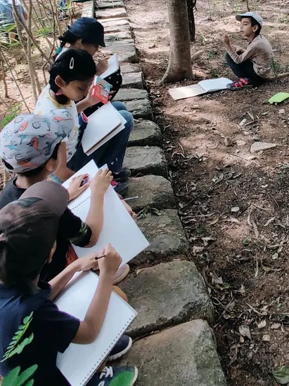 Children sit along a stone path, deeply engaged in sketching and writing in their nature journals during a park visit.
