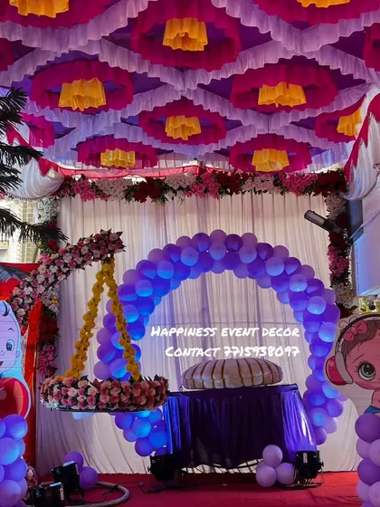 This naming ceremony decor features a beautiful tent-style ceiling with pink and yellow drapes. The stage has a flower-decorated cradle and a purple balloon arch, creating a festive look.
