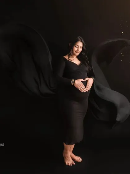 A dramatic fine art maternity portrait of a woman in a sleek black gown against a dark background. The fabric flows out like wings, and she makes a heart shape with her hands over her baby bump, creating a moody and artistic pregnancy photo.