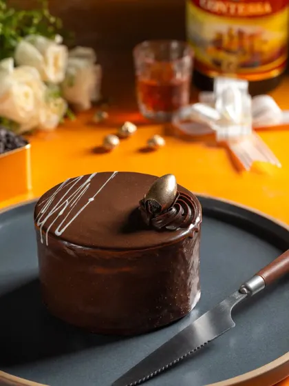 A rich chocolate truffle mini cake, styled with a liquor bottle and white roses to suggest a sophisticated flavor pairing.