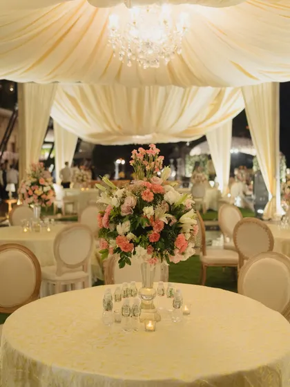An elegant outdoor dinner setting under a draped canopy with a crystal chandelier. The soft cream and pink floral centerpieces and classic round-back chairs create a sophisticated and intimate atmosphere perfect for a formal corporate dinner.
