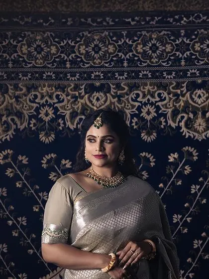 A wide shot of a model in a shimmering silver saree, posed against an ornate blue and gold carpet. The dramatic lighting and unique backdrop create a regal and theatrical portrait.