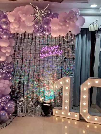 A glamorous 10th birthday setup with a silver shimmer wall. The decor includes a purple balloon arch, a neon sign, and large light-up marquee numbers.