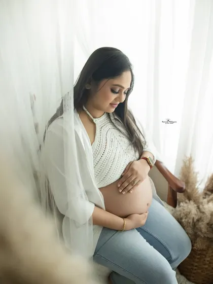 A serene and modern maternity portrait. The mother-to-be is dressed in a casual crochet top and jeans, sitting by a window for soft, beautiful light.