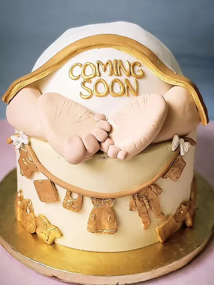 A cute "Coming Soon" baby shower cake. The design features tiny fondant baby feet and a clothesline with baby clothes, all in a classic white and gold theme.