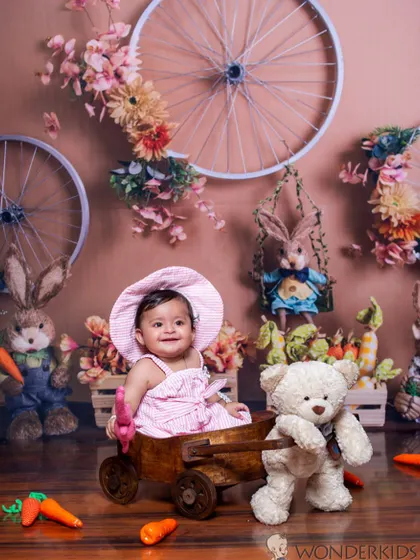 A happy baby girl in a pink gingham dress sits in a wooden cart, surrounded by an adorable Easter or spring-themed setup with bunnies, carrots, and flowers.