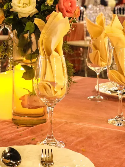 A close-up of a table centerpiece with candles and flowers. The yellow folded napkins in the wine glasses add a pop of color that complements the warm candlelight.